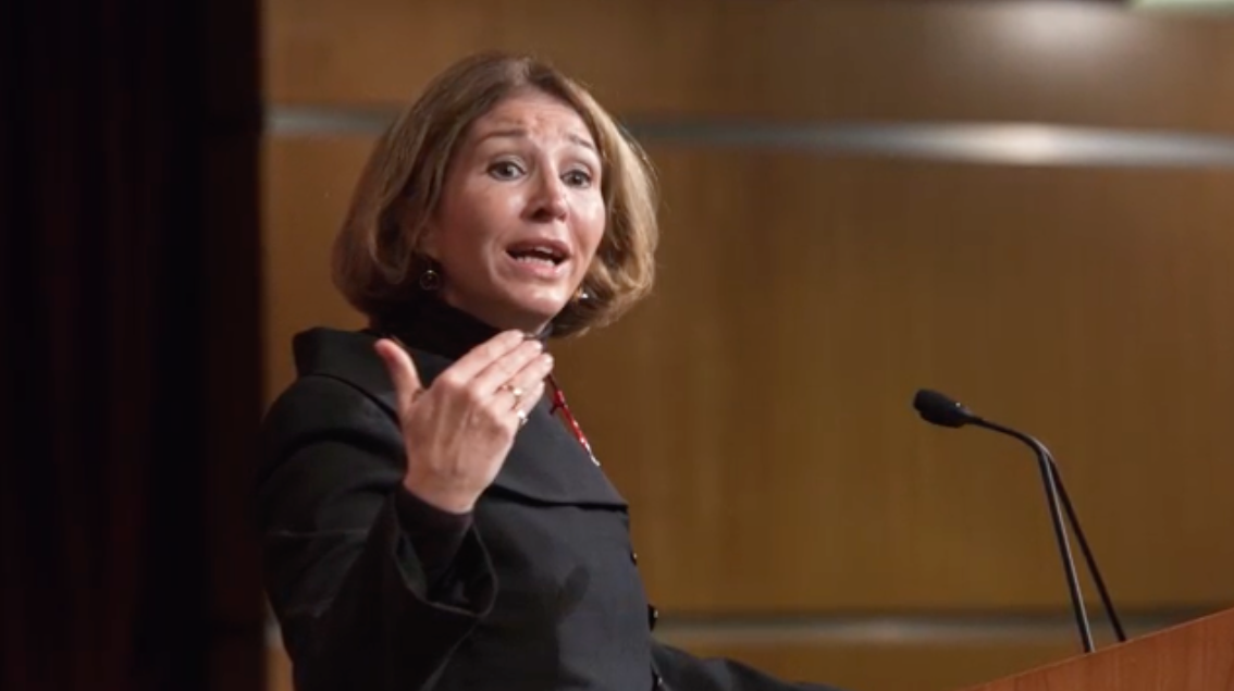 Anne-Marie Slaughter, a light skinned woman with chin-length light brown hair, speaks at a podium and gestures with her right hand.
