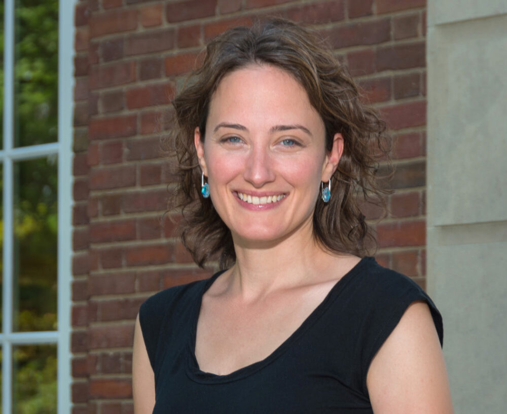 Elizabeth Hansen, a light skinned woman with shoulder length brown hair, stands in front of a brick wall wearing a sleeveless black top.