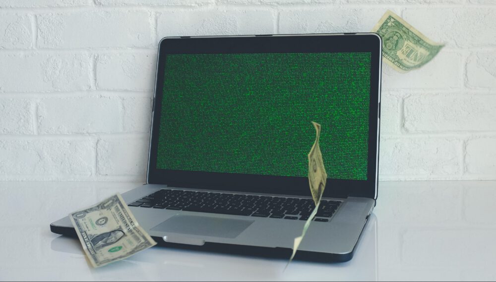image of a laptop computer with dollar bills falling around it, symbolizing profit from technology. Photo by NeONBRAND on Unsplash.