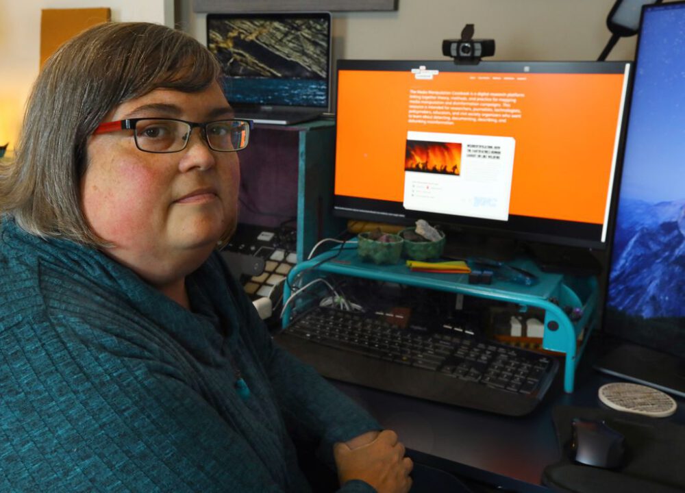 Photograph of Joan Donovan, a white woman, at her desk looking at a computer screen