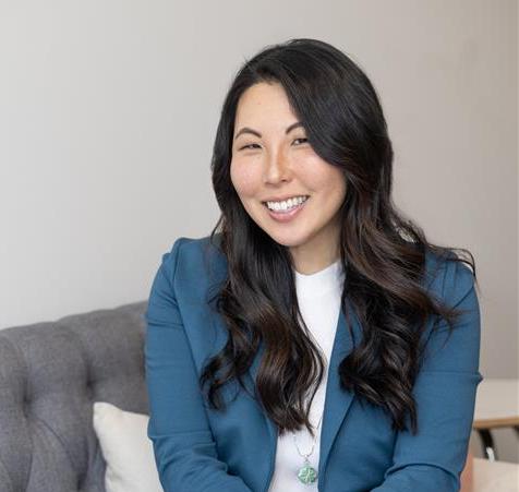 Laura Manley, a woman with long wavy dark hair, sits on a gray couch while wearing a teal blazer and white shirt.
