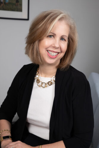 Nancy Gibbs, a light skinned woman with chin-length blond hair, smiles into the camera wearing a black sweater, white shirt, and gold necklace.
