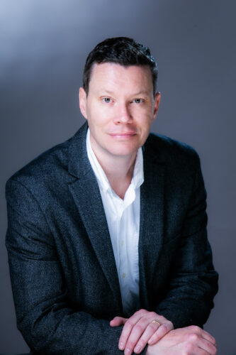 Clark Merrefield headshot. Alt text: Studio portrait of a man in a dark blazer and white shirt, seated against a gray background, looking at the camera with a calm expression.