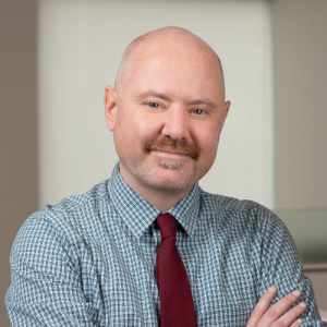 Photo of josh shank, a light-skinned man with a mustache wearing a blue shirt and red tie