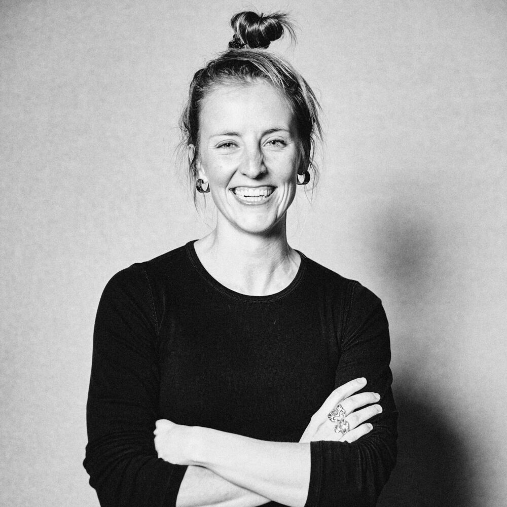 black and white photo of Gabby Malina, a light-skinned woman with her hair in a bun, wearing a dark colored shirt, smiling with her arms crossed