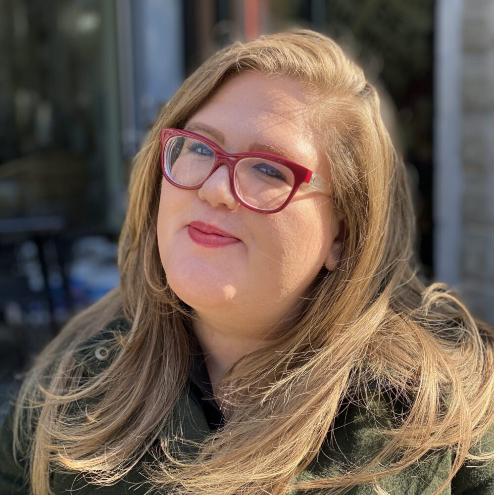 photo showing Megan O'Neil, a light skinned woman with long blonde hair, smiling at the camera, wearing red glasses