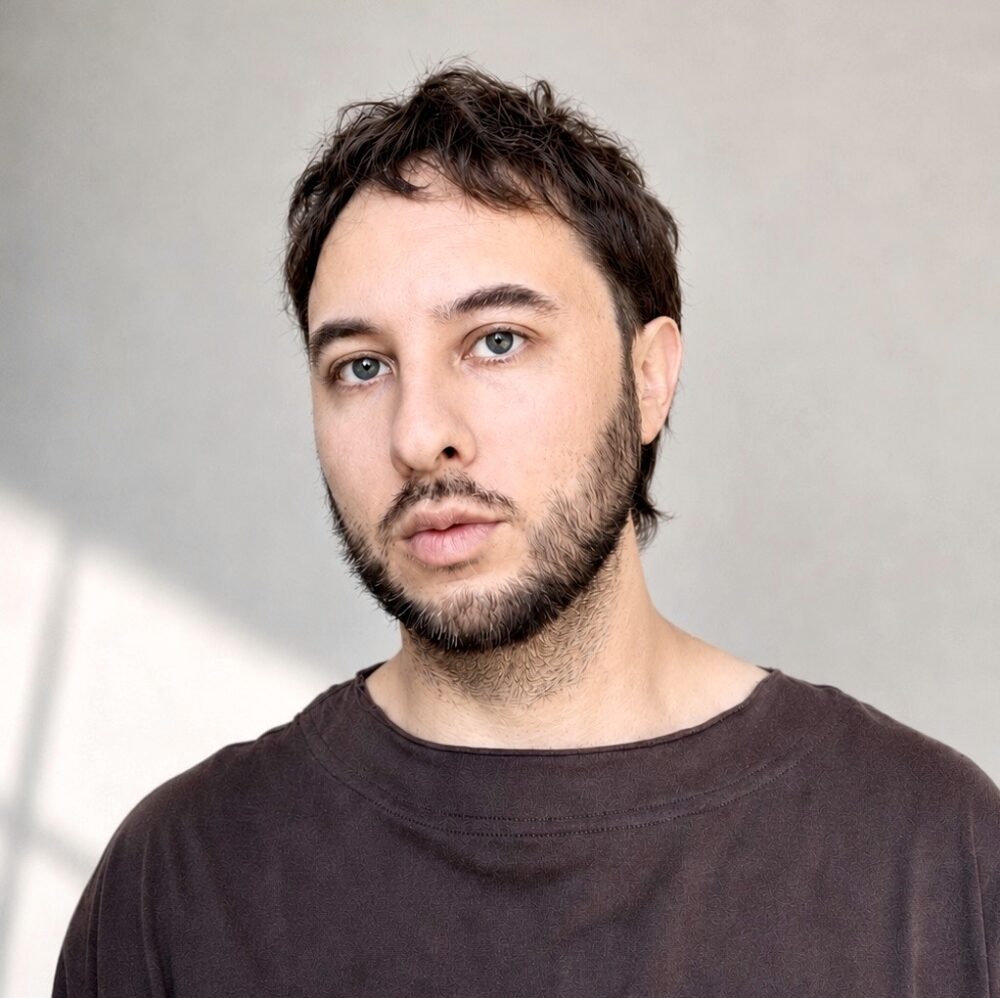 Enrique Pedraza-Botero, a light skinned man with short brown hair and short facial hair, wearing a brown shirt standing in front of a beige wall with light from a window hitting the bottom left corner.