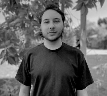 A man with dark hair standing in front of some trees and grass wearing a dark colored t-shirt.