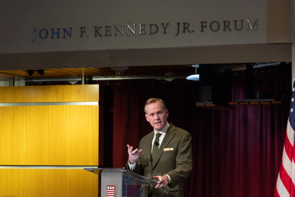 John Dickerson, a light skinned man with light reddish colored hair, speaks at a podium below a sign that say s