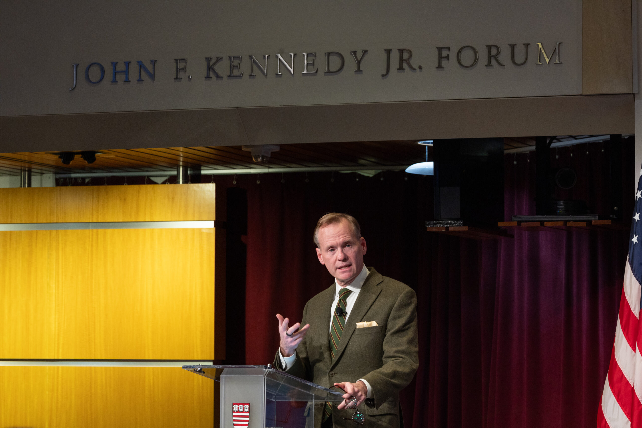 John Dickerson, a light skinned man with light reddish colored hair, speaks at a podium below a sign that say s"John F. Kennedy Jr. Forum" wearing a green jacket, white shirt, and tie.