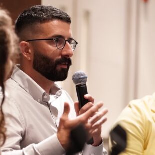 A man with dark hair and a beard, wearing glasses and a white button-down shirt, shown from the side, speaks into a microphone and gestures with his hand