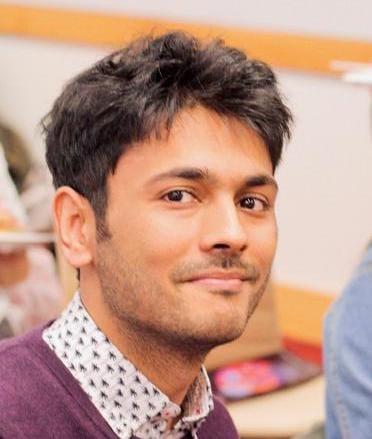 Rehan Mirza, a medium skinned man with short dark hair, sitting in an indoor space wearing a purple sweater with collared shirt