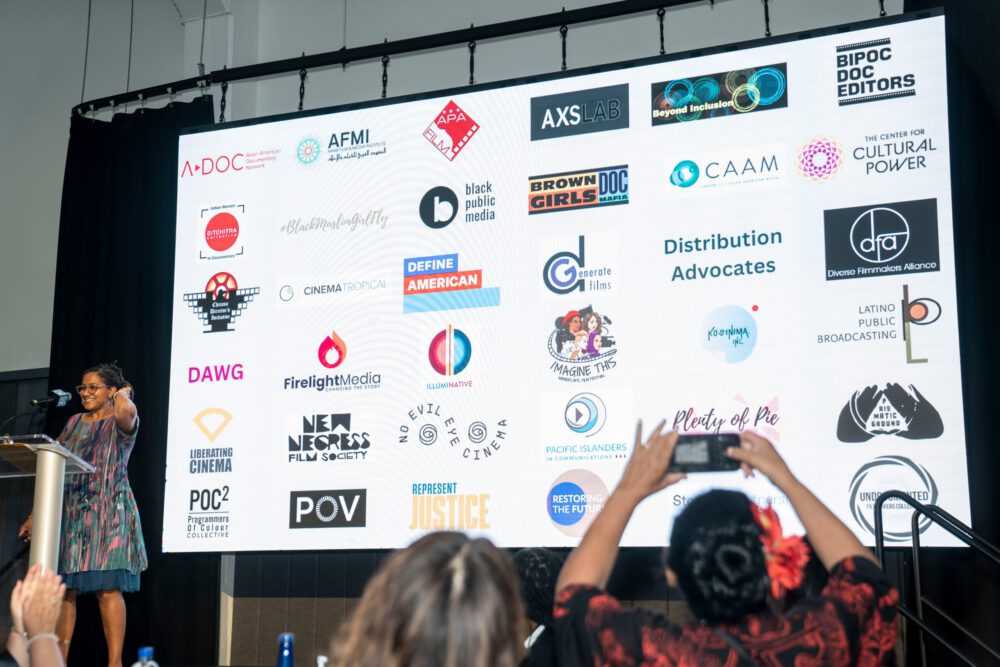 Photograph of a person standing in front of a screen filled with organization logos
