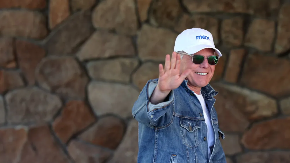 Photograph of a person wearing a white hat and sunglasses waving one arm forward