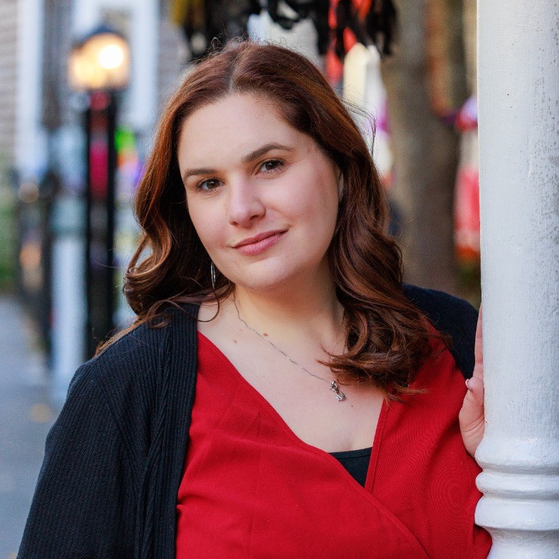 Natalie Solomon, a woman with wavy brown hair, wearing a red top and black cardigan, poses outdoors near a white pillar. The background is softly blurred with street elements.