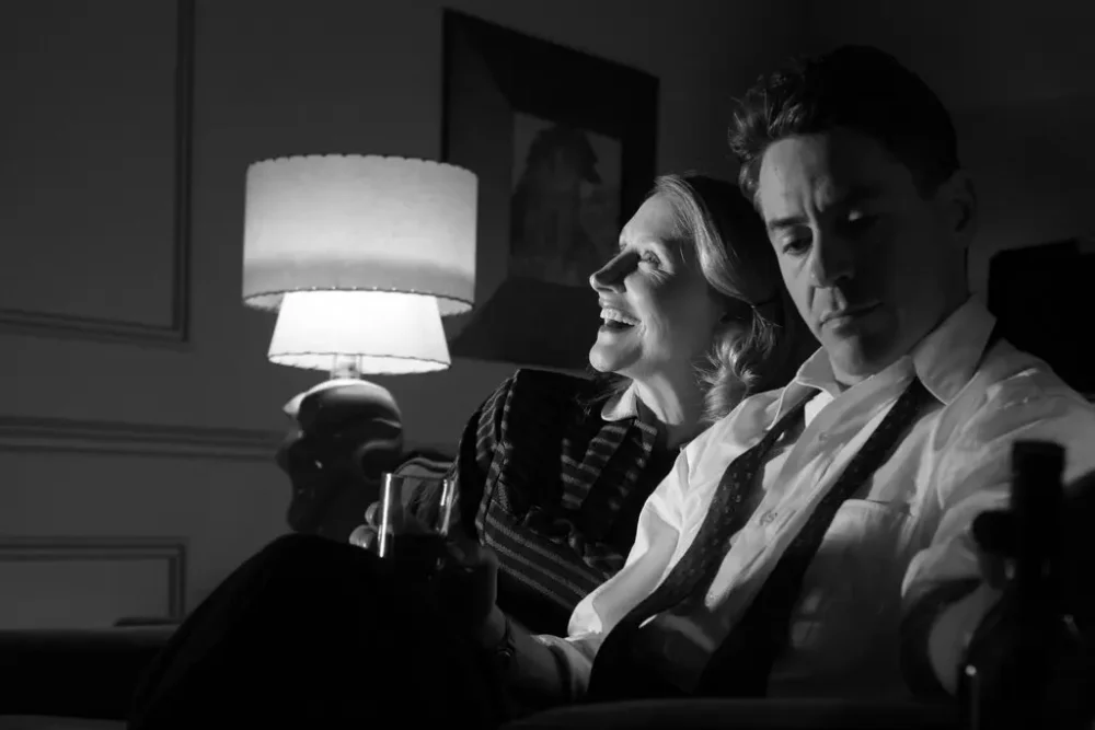 Black and white photograph of two people, one smiling and looking upward and one looking seriously downward, sitting next to a lamp