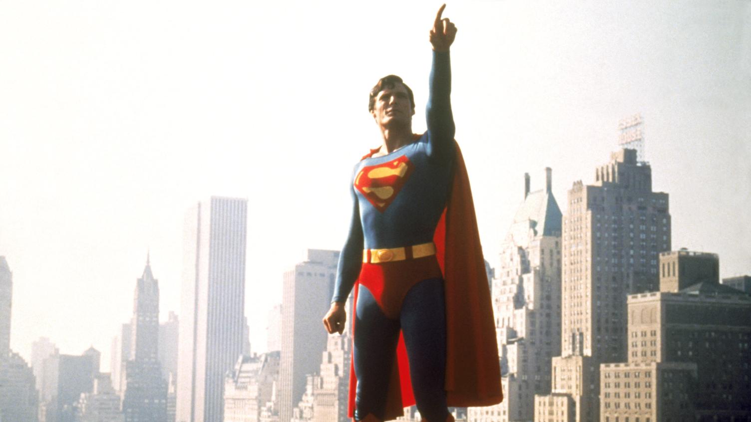 Photograph of person wearing a blue Superman costume with a red cape pointing up to the sky