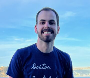 Evangelos Kassos, a light-skinned man with short trimmed hair and facial hair wearing a t-shirt stands in front of water and a blue sky.