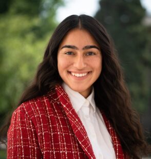 Muskan Walia, a medium skinned woman with long dark hair smiles wearing a red blazer and white shirt stands outside.
