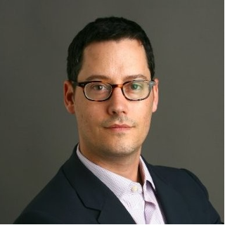 photo of Ben Monnie, a light skinned man with short dark hair wearing glasses and a button down shirt with a dark suit jacket.