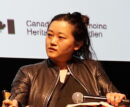 Abby Sun, a woman of Asian descent with dark hair pulled back, sits on a stage with a microphone, wearing a dark leather jacket