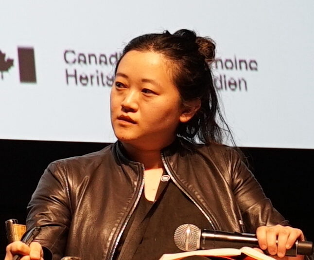 Abby Sun, a woman of Asian descent with dark hair pulled back, sits on a stage with a microphone, wearing a dark leather jacket