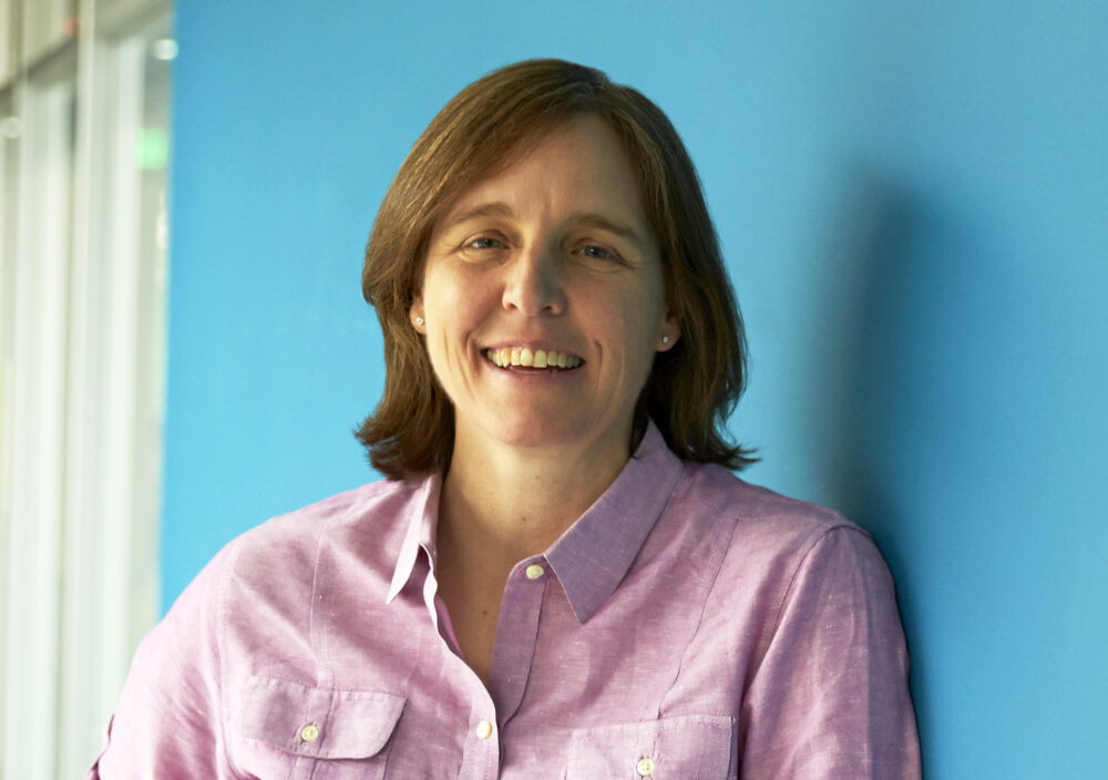 Megan Smith, a woman with shoulder length hair wearing a pink button down shirt, stands against a blue wall.