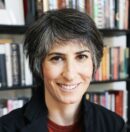 a light skinned woman with short dark hair wearing a black jacket and red shirt, sitting in front of a bookshelf, smiles into the camera