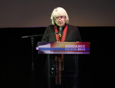 Mary Lampson, a light skinned woman with chin length gray hair, standing at a lecturn