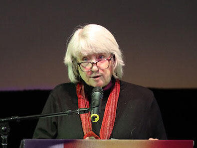 Mary Lampson, a light skinned woman with chin length gray hair, standing at a lecturn