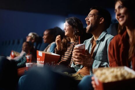 People in movie theatre seats with drinks and popcorn in hand smiling up at the screen