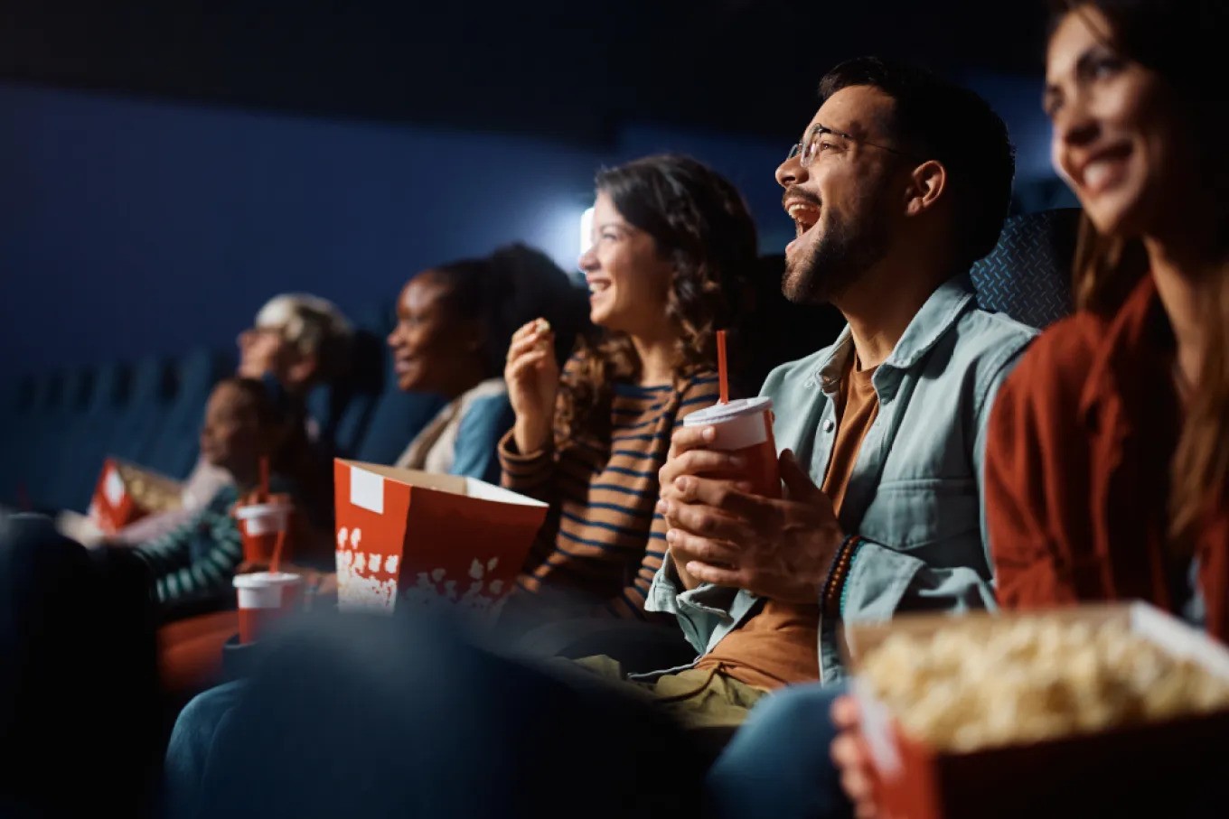 People in movie theatre seats with drinks and popcorn in hand smiling up at the screen