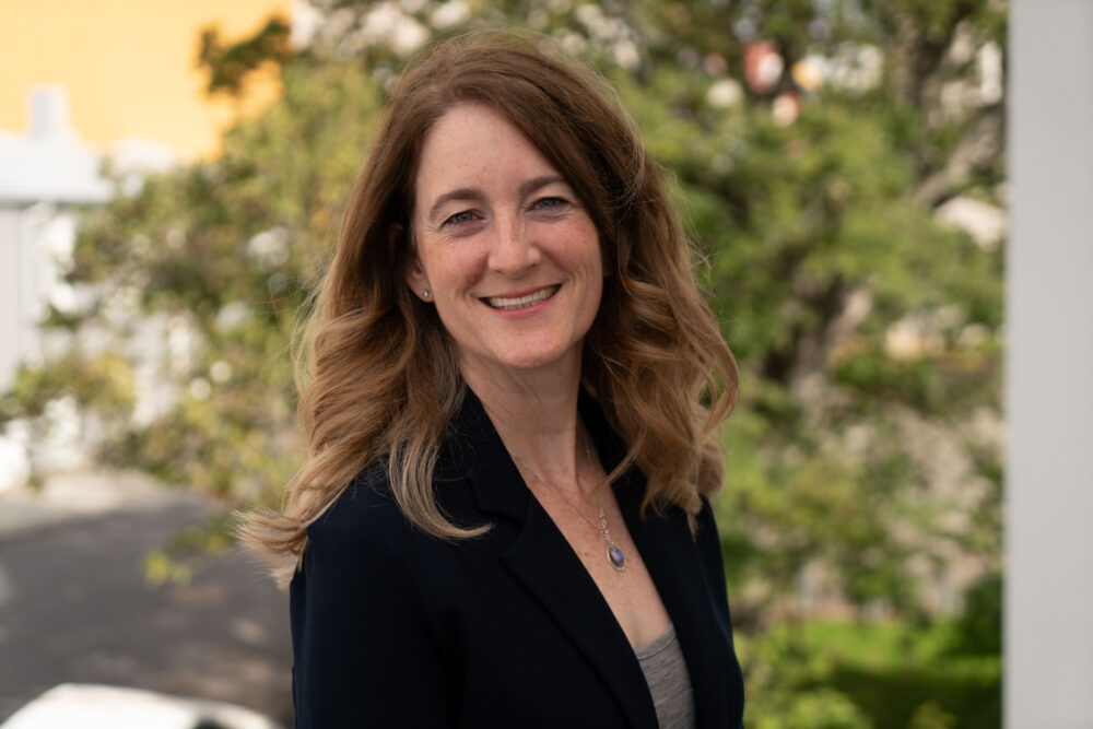 Candace Bertotti, a woman with long red hair wearing a black blazer stands outside and looks over her shoulder at the camera.