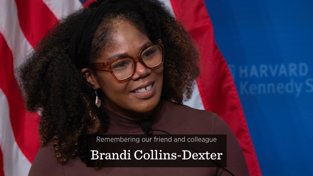 Brandi Collins-Dexter, a Black woman wearing glasses and a brown shirt, smiles on stage at the JFK Jr. Forum at Harvard Kennedy School. The words 