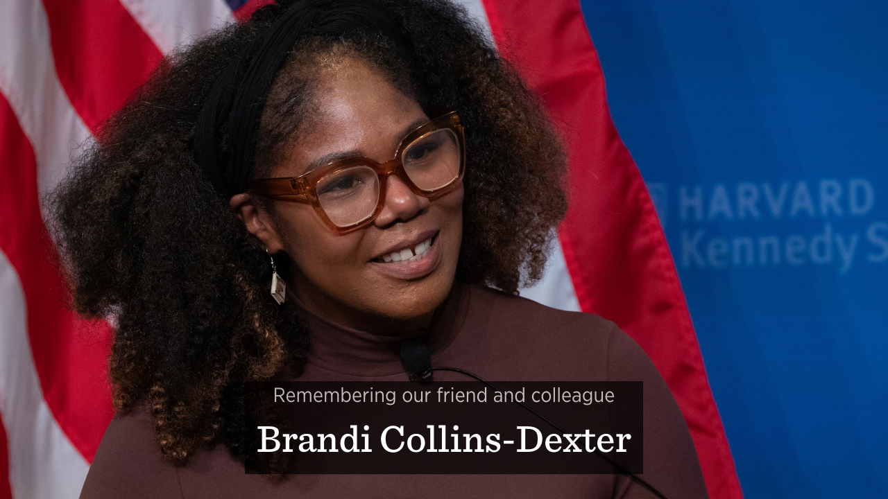 Brandi Collins-Dexter, a Black woman wearing glasses and a brown shirt, smiles on stage at the JFK Jr. Forum at Harvard Kennedy School. The words "Remembering our friend and colleague Brandi Collins-Dexter" are superimposed over the image.