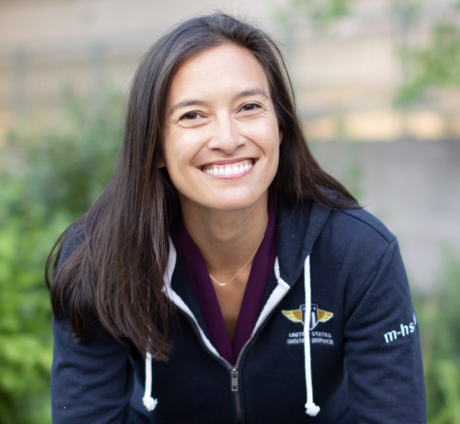 Mina Hsiang, a woman of Asian descent with light skin and long dark hair, smiles towards the camera wearing a blue zip-up sweatshirt with the USDS logo on the front.