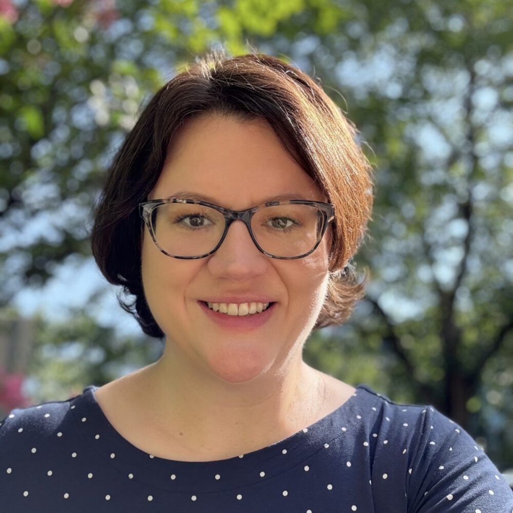 Rebecca Piazza, a light skinned woman with short reddish brown hear wearing glasses and a blue shirt sitting outside near trees