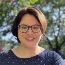 Rebecca Piazza, a light skinned woman with short reddish brown hear wearing glasses and a blue shirt sitting outside near trees