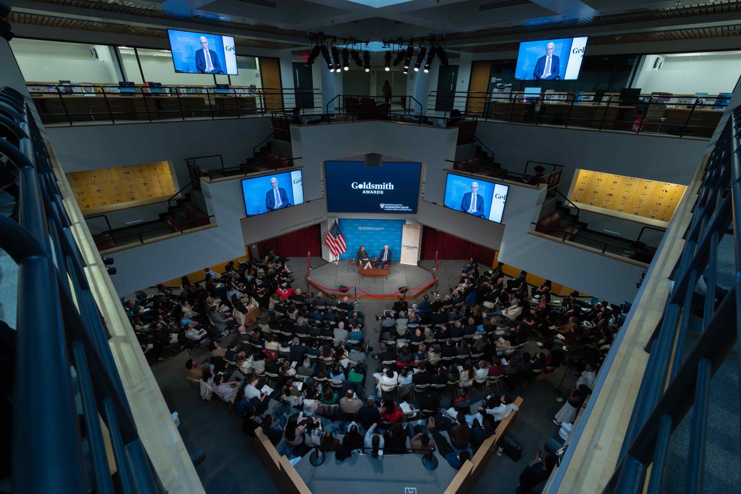 The JFK Jr Forum at Harvard Kennedy School, a large diamond-shaped multi-story indoor space with seats on the floor level and large screens displaying the stage at each level.
