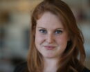 Allison Abbott, a light skinned woman with red hair and blue eyes, looks into the camera with a blurred background behind her