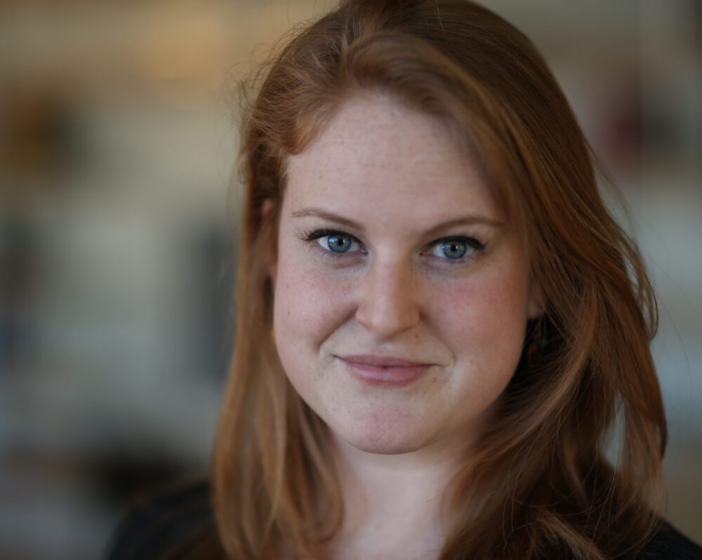 Allison Abbott, a light skinned woman with red hair and blue eyes, looks into the camera with a blurred background behind her