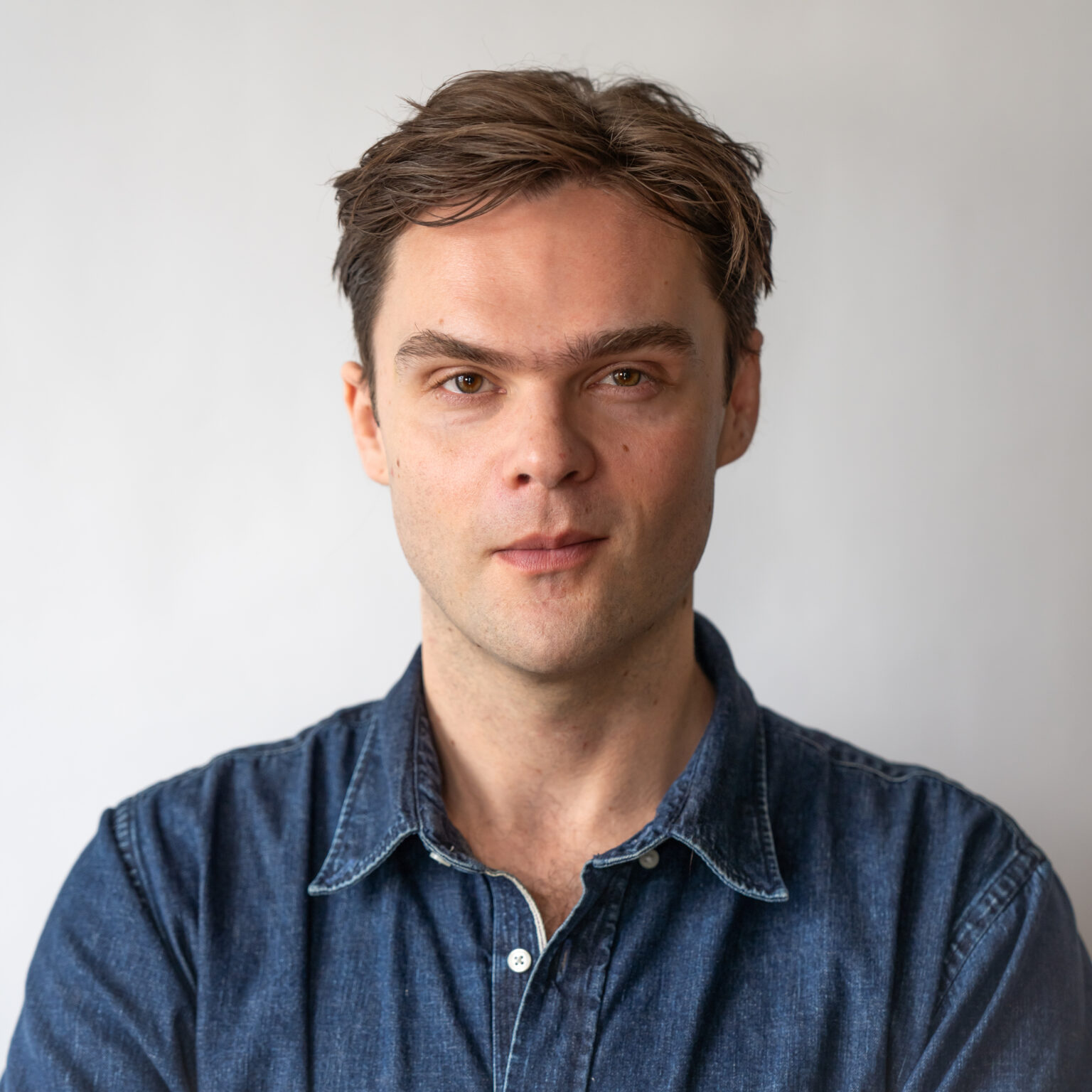 Ben Reninga, a light skinned man with short brown hair wearing a dark blue button down shirt faces the camara with a white background behind him.