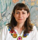 Erin Zimmer Strenio, a light skinned woman with shoulder length brown hair wears a white shirt with embroidered flowers, in front of a decorative mural.