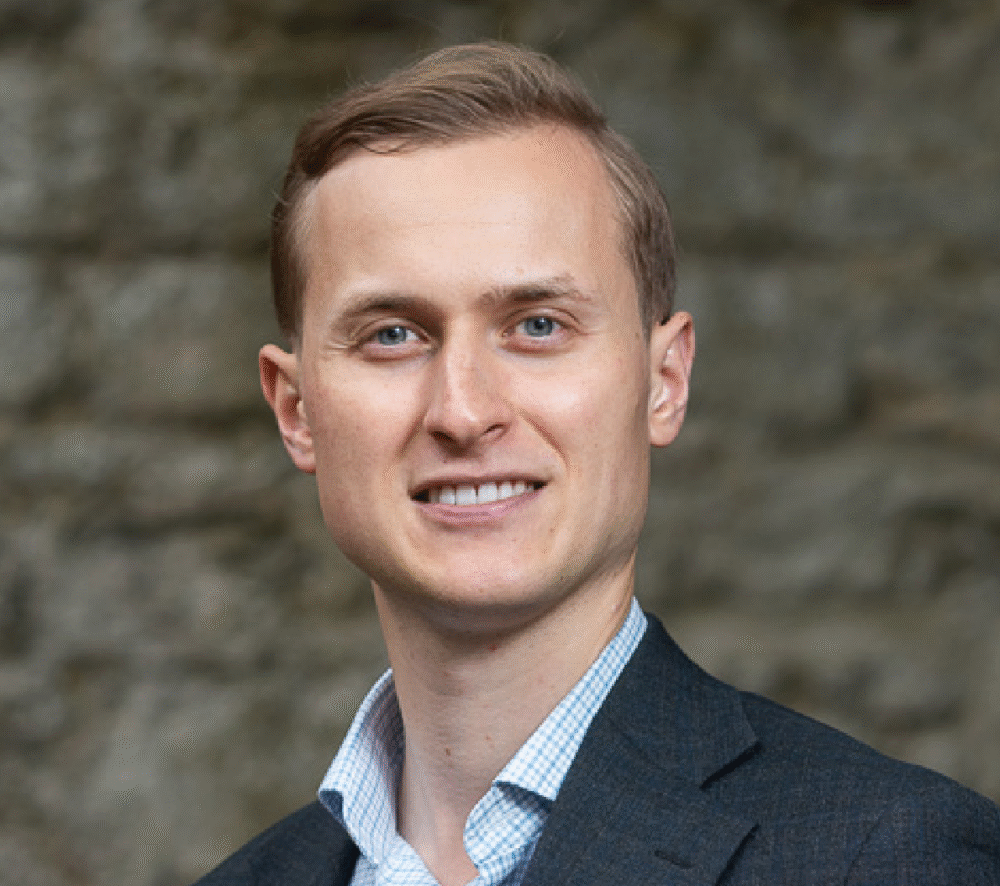 Dr. Rakoen Maertens, a light skinned man with short light brown hair wearing a shirt and jacket, stands in front of a stone wall