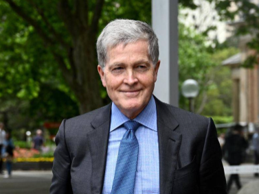 Steven Johnson, a light skinned man with short gray hair wearing a blue suit stands outside near some trees and a building
