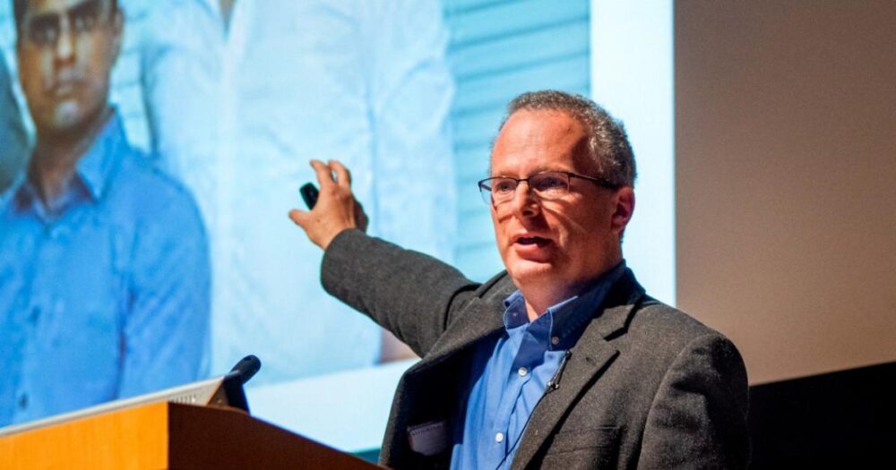David Lazer, a man in glasses and a suit jacket is giving a presentation, gesturing toward a projected image behind him. He is standing at a podium and speaking to an audience.