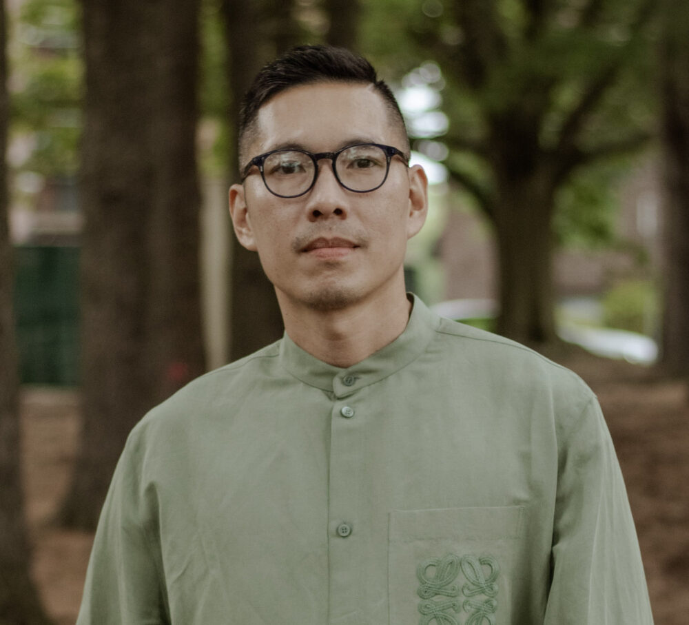 Jonathan Corpus Ong, a man with short dark hair wearing black framed glasses and a green button down shirt, stands in a grove of trees