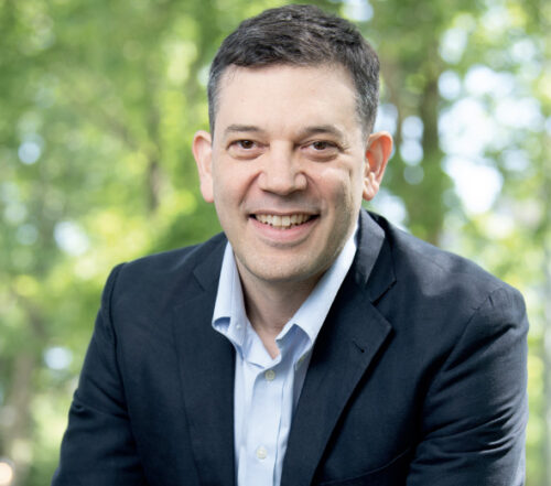 Man with short dark hair wearing a navy jacket and light blue collared shirt, smiling at the camera. The background is outdoors with green trees and soft natural light.