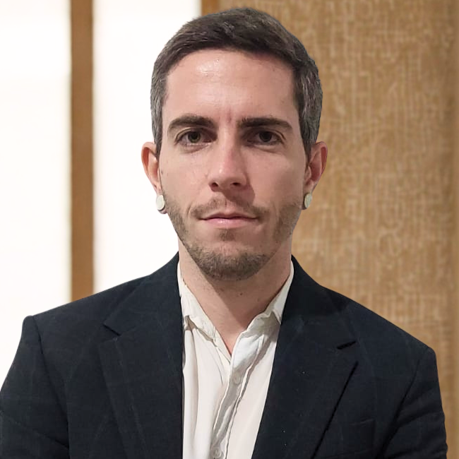 Photo of Thales Lelo, a man with short dark hair and a serious expression, wearing a white shirt with a dark jacket and flat round white earrings, standing in front of a neutral white and brown wall.