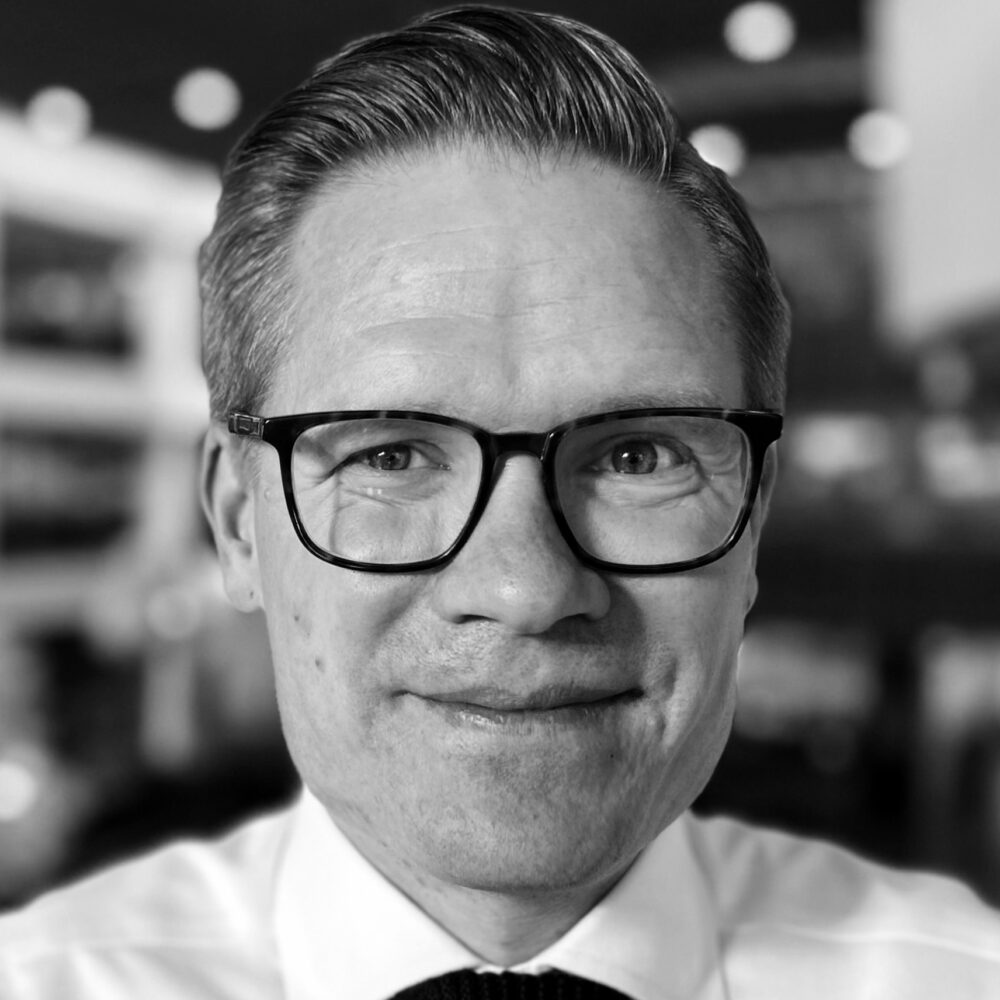 Rasmus Kleis Nielsen, a man with short hair and glasses wearing a white collared shirt and tie, smiles into the camera in a black and white photo